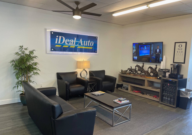 Modern car dealership lounge with black chairs, TV, coffee table, lamp, and iDeal Auto sign | iDeal Auto Service