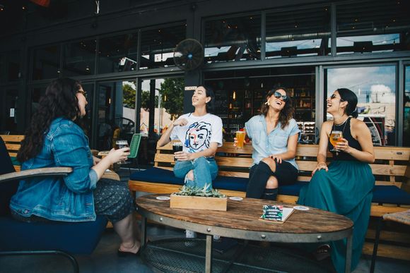 Group of friends sitting outside a café or brewery patio, laughing and chatting while holding drinks around a wooden table.