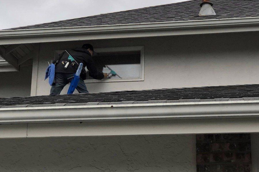 Worker on a roof cleaning or inspecting a second-story window above a white house exterior