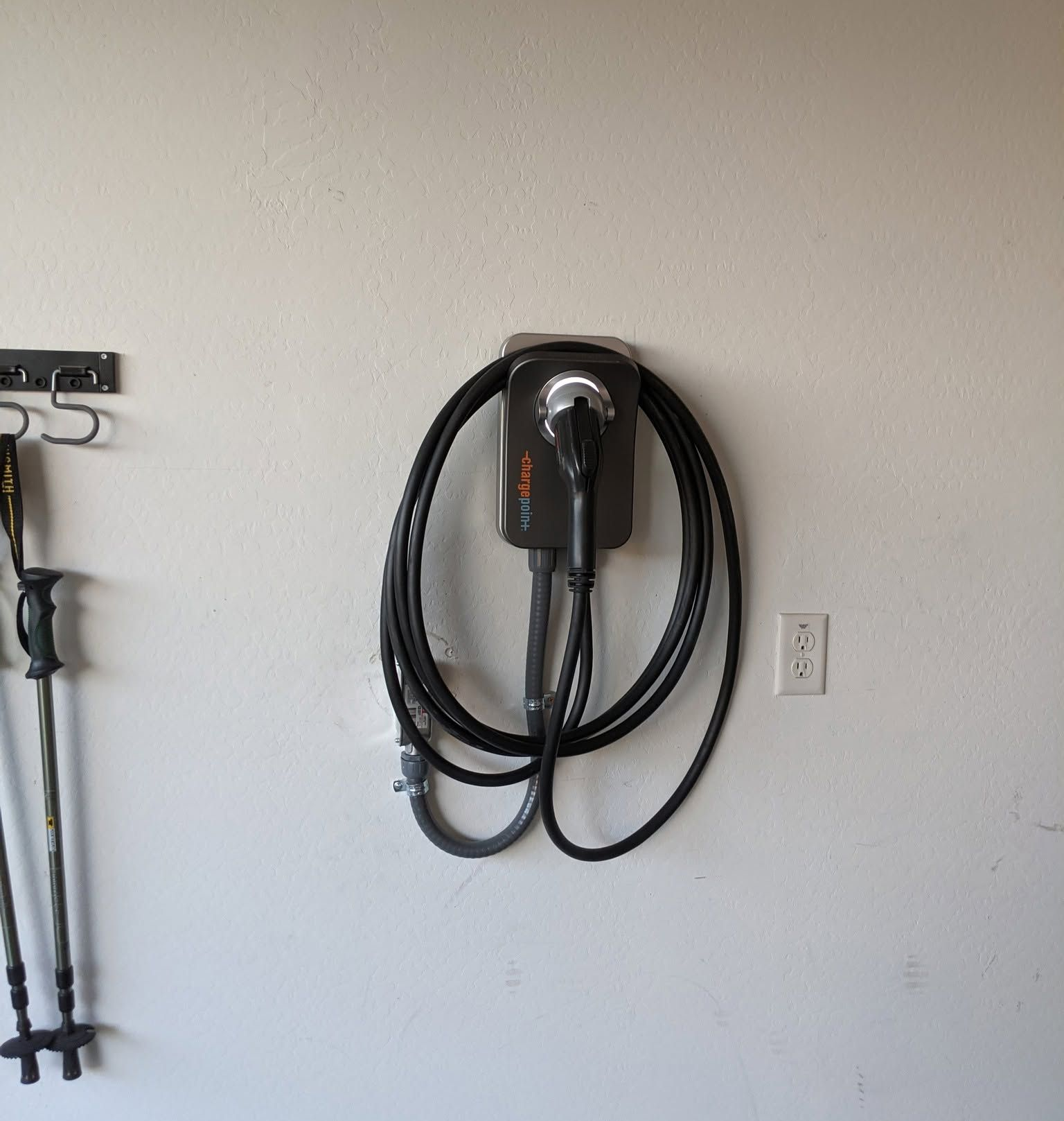 Wall-mounted EV charger with coiled black cable beside a power outlet on a white garage wall