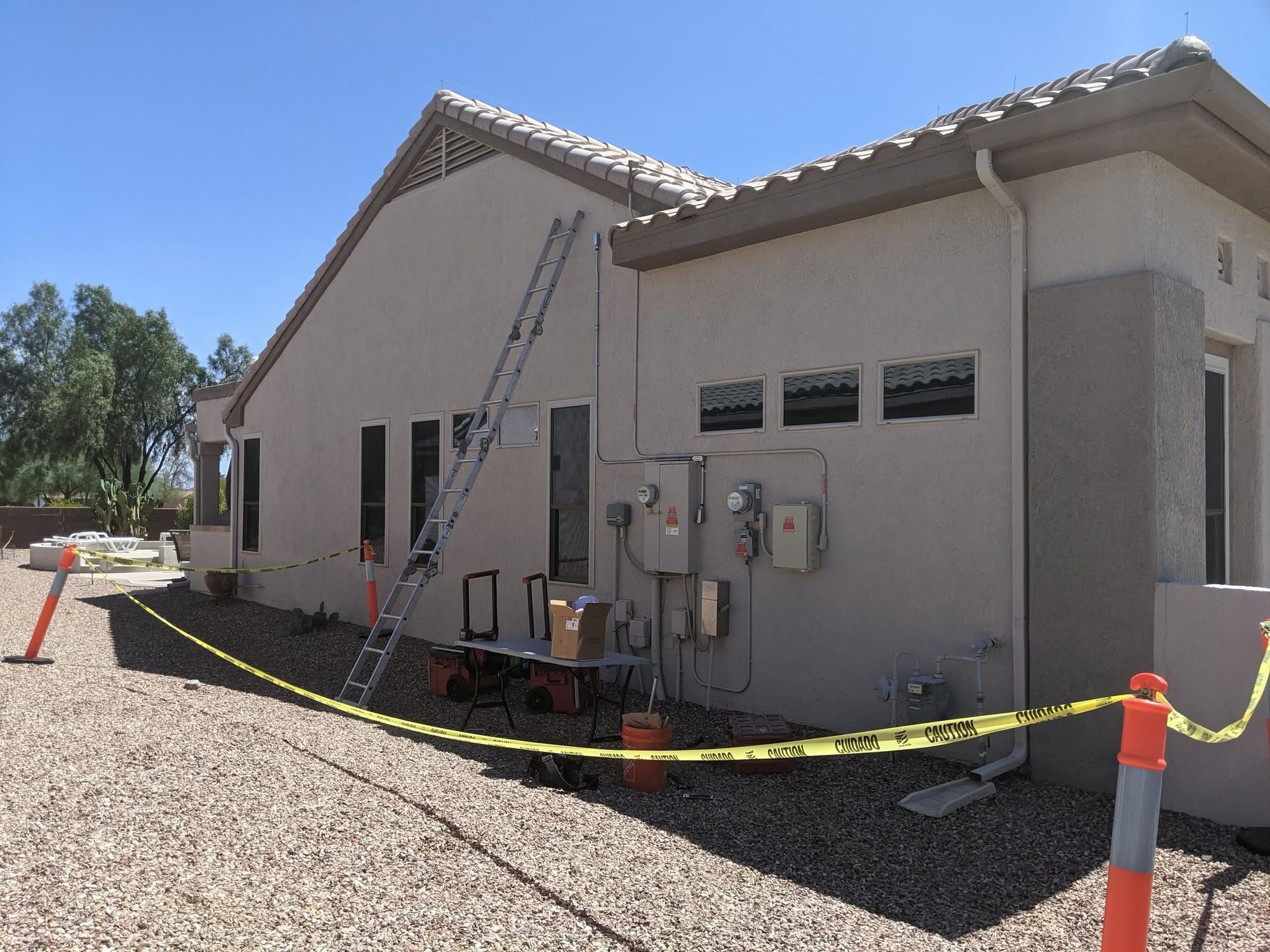 Side view of a small utility building with electrical meters, ladder, and caution tape around the gravel lot