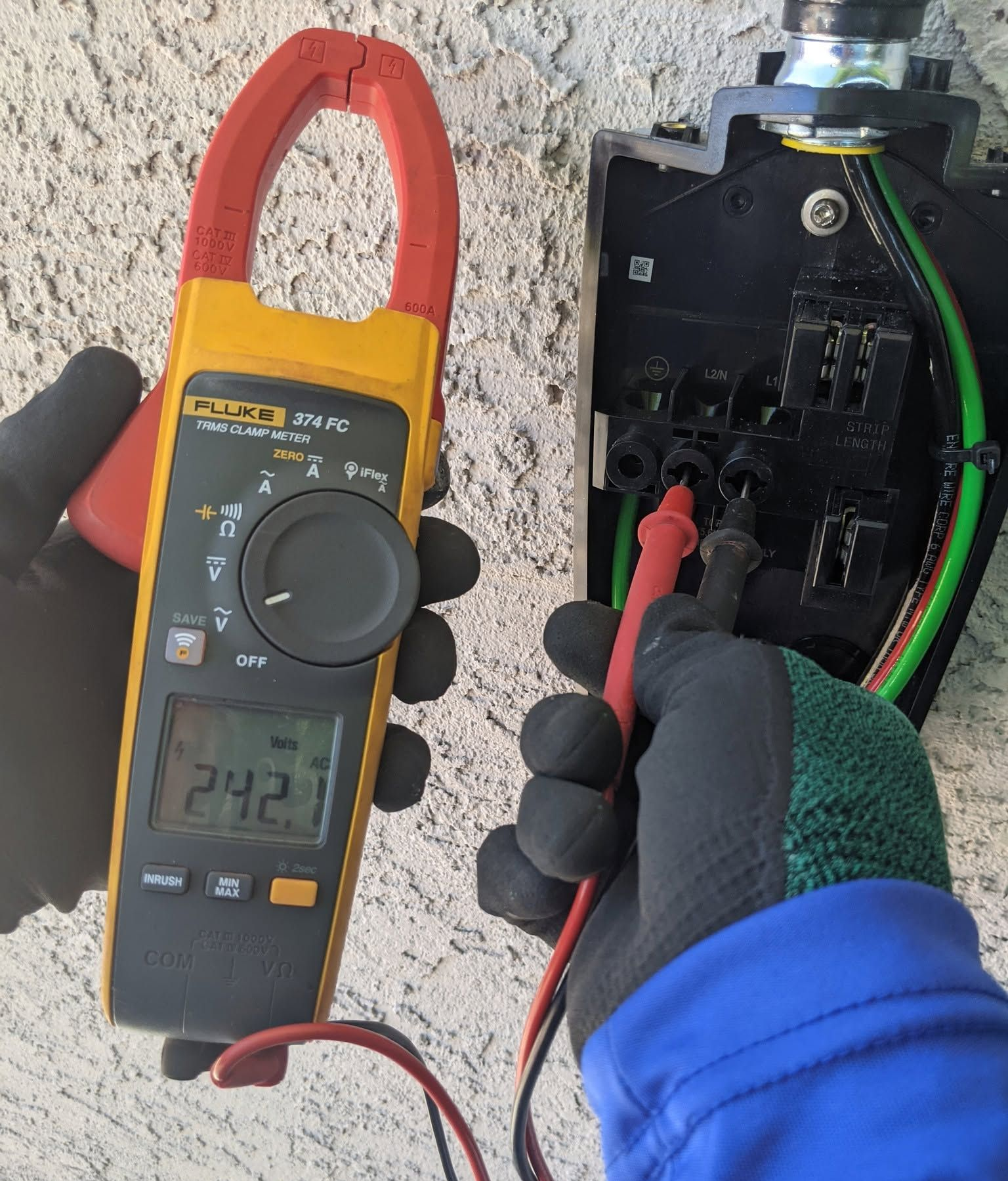 Gloved hand testing an electrical panel with a clamp meter and red probes outdoors on a wall.