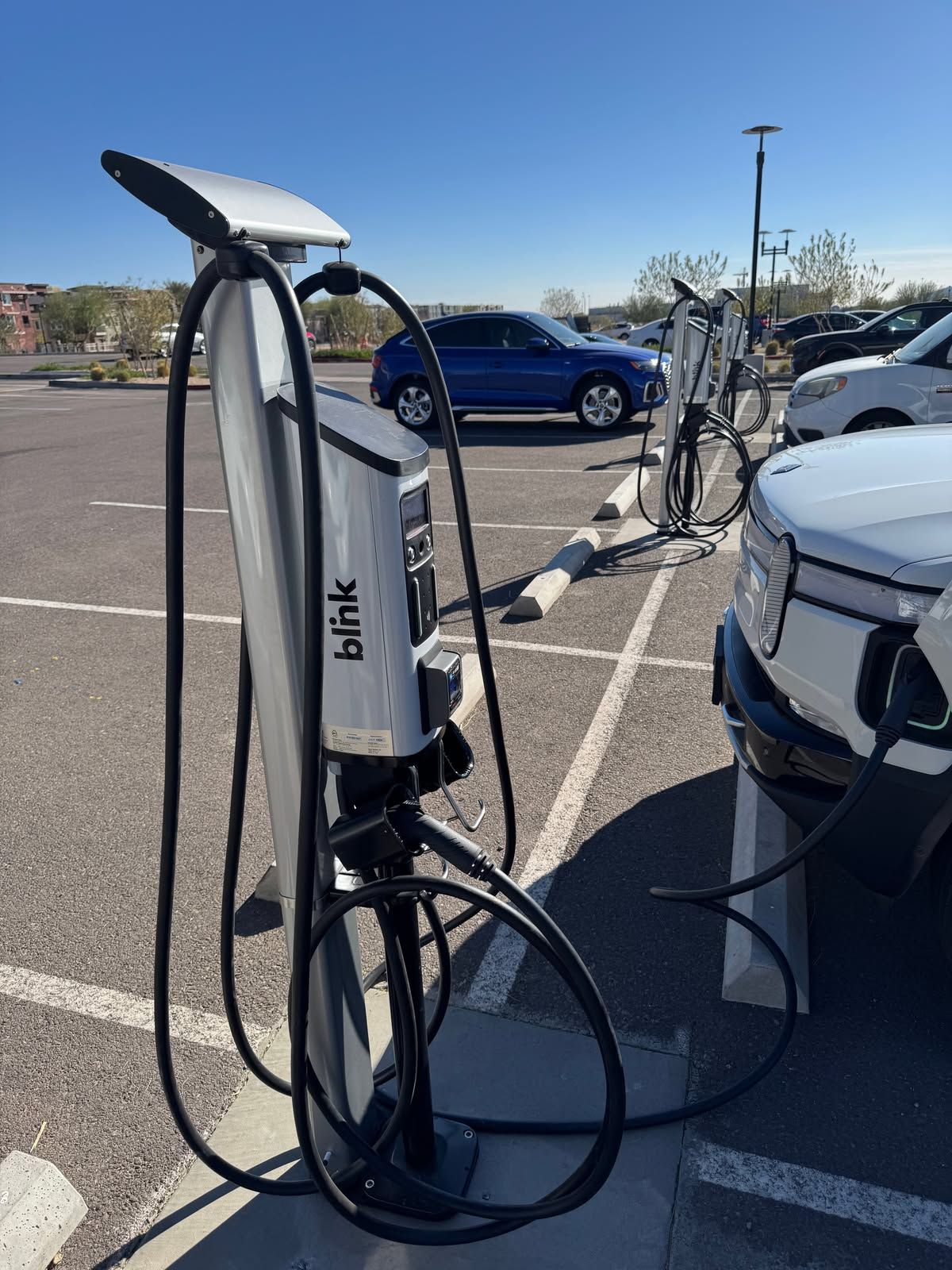 Public EV charging station with a white charger and black cables in a parking lot, blue car nearby.