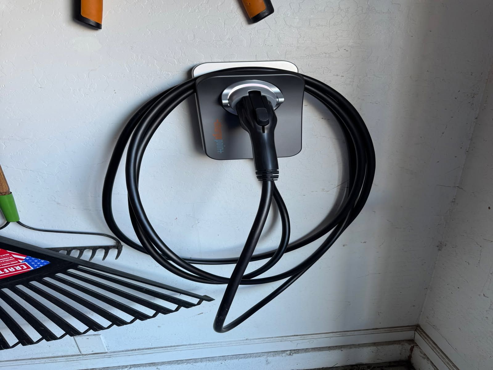 Black charging cable coiled on a wall-mounted outlet beside a rake handle and broom bristles.