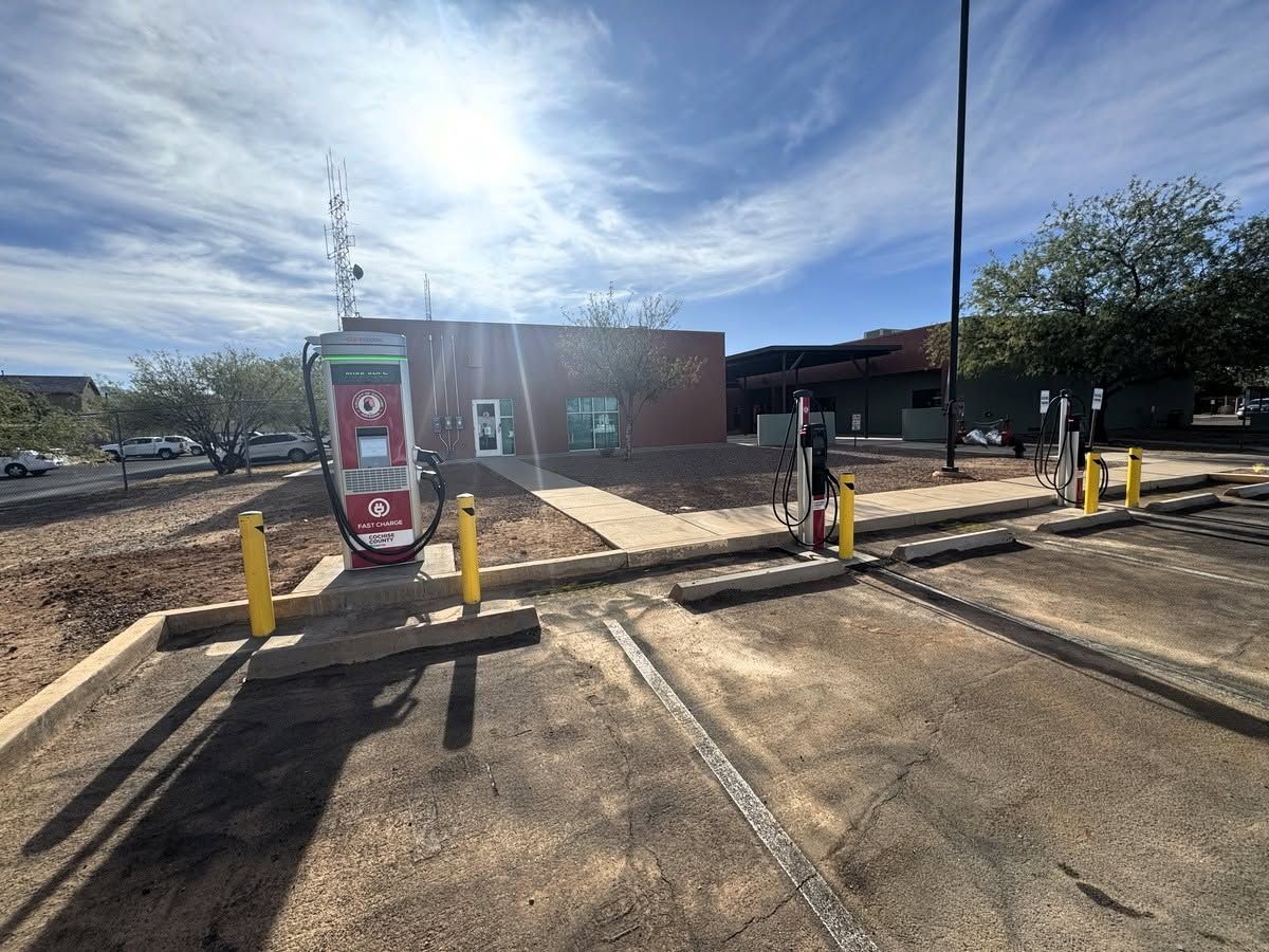Sunlit parking lot with EV charging stations, bollards, and a brick building under a cloudy sky