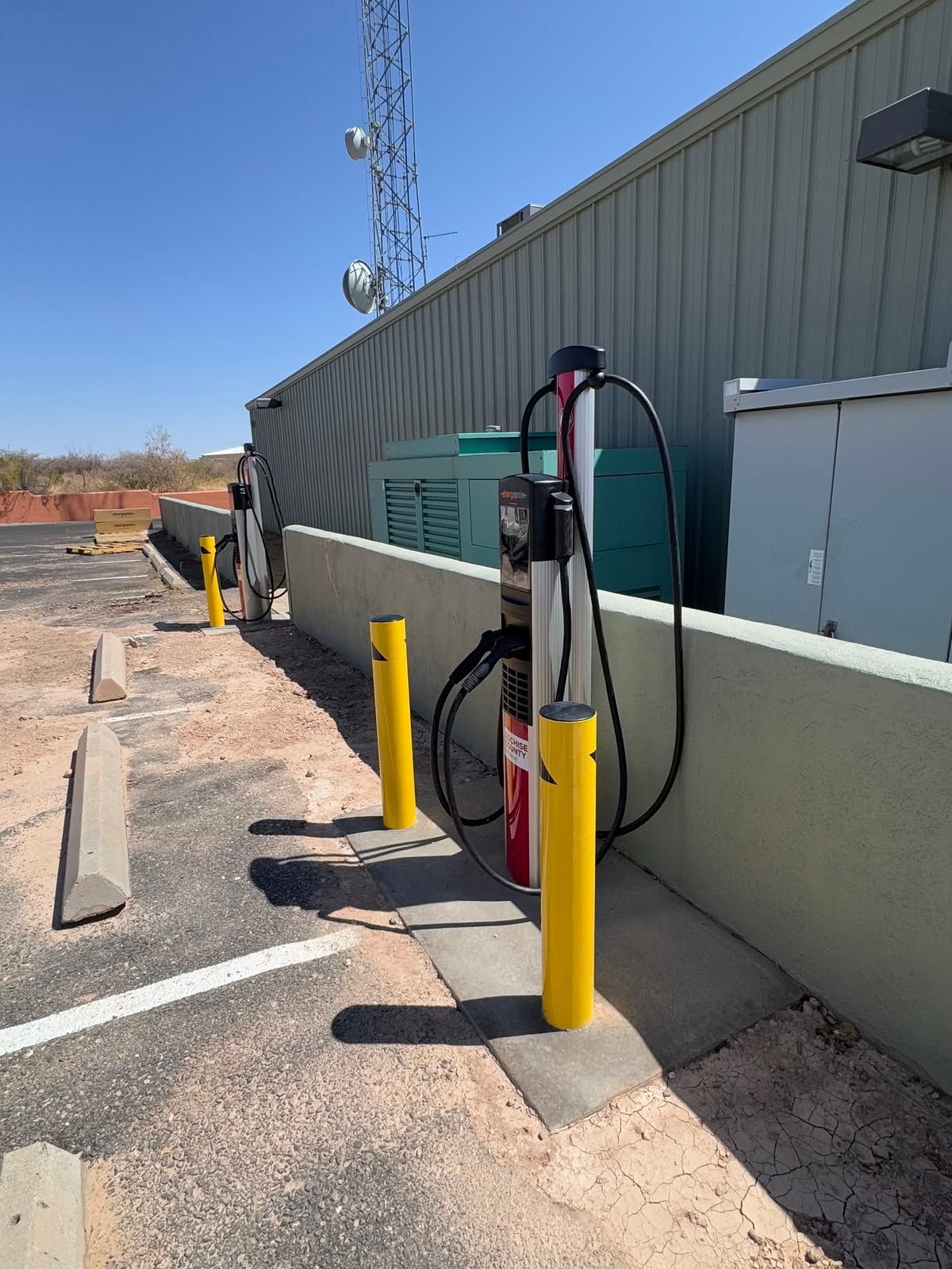 Electric vehicle charging station beside a gray building, with yellow bollards and a red fire extinguisher.