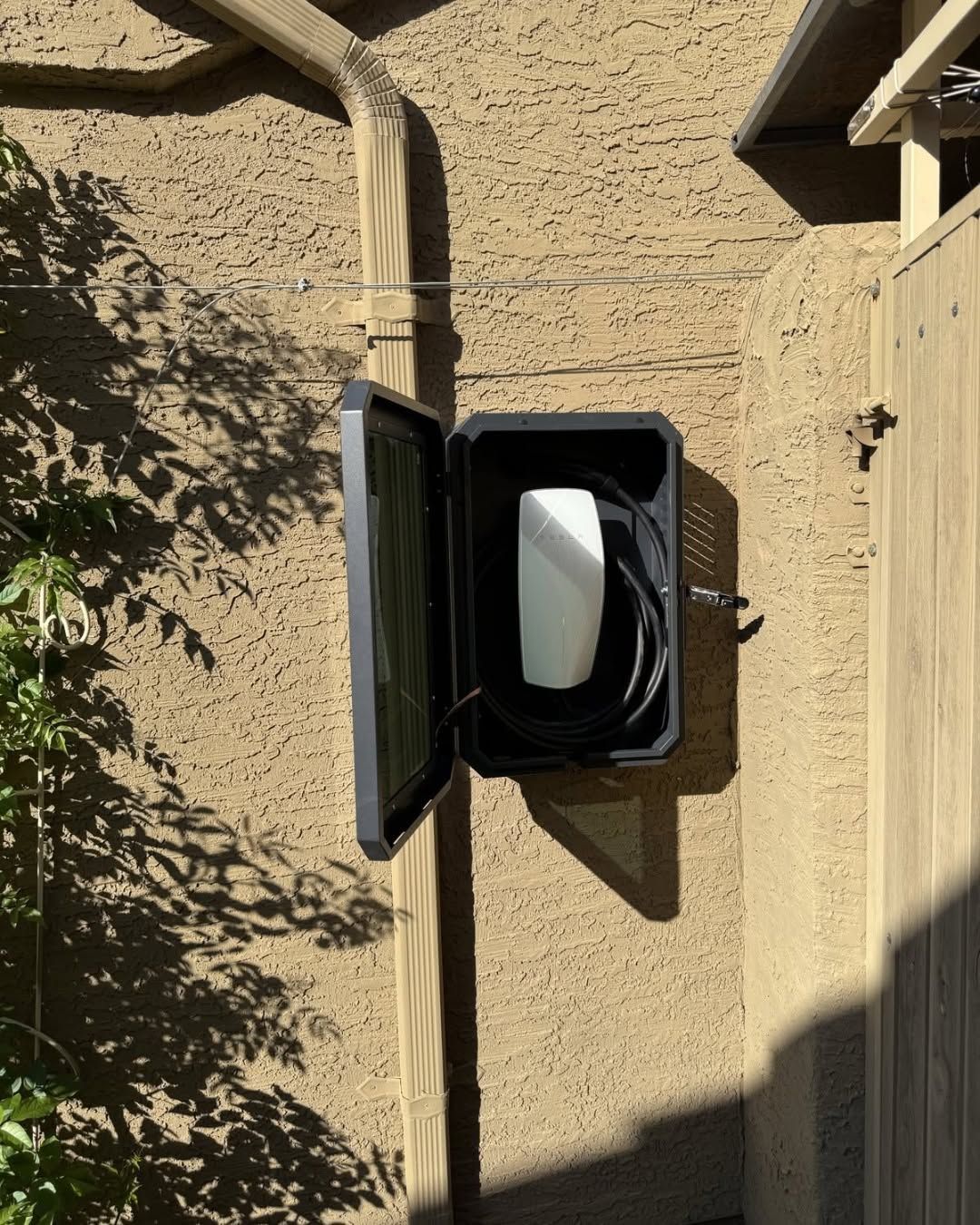 Gray outdoor electrical panel mounted on a beige stucco wall beside a wooden gate.
