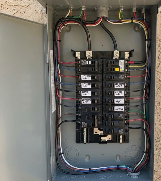 Open electrical breaker panel with labeled circuits and multicolored wires inside a gray cabinet