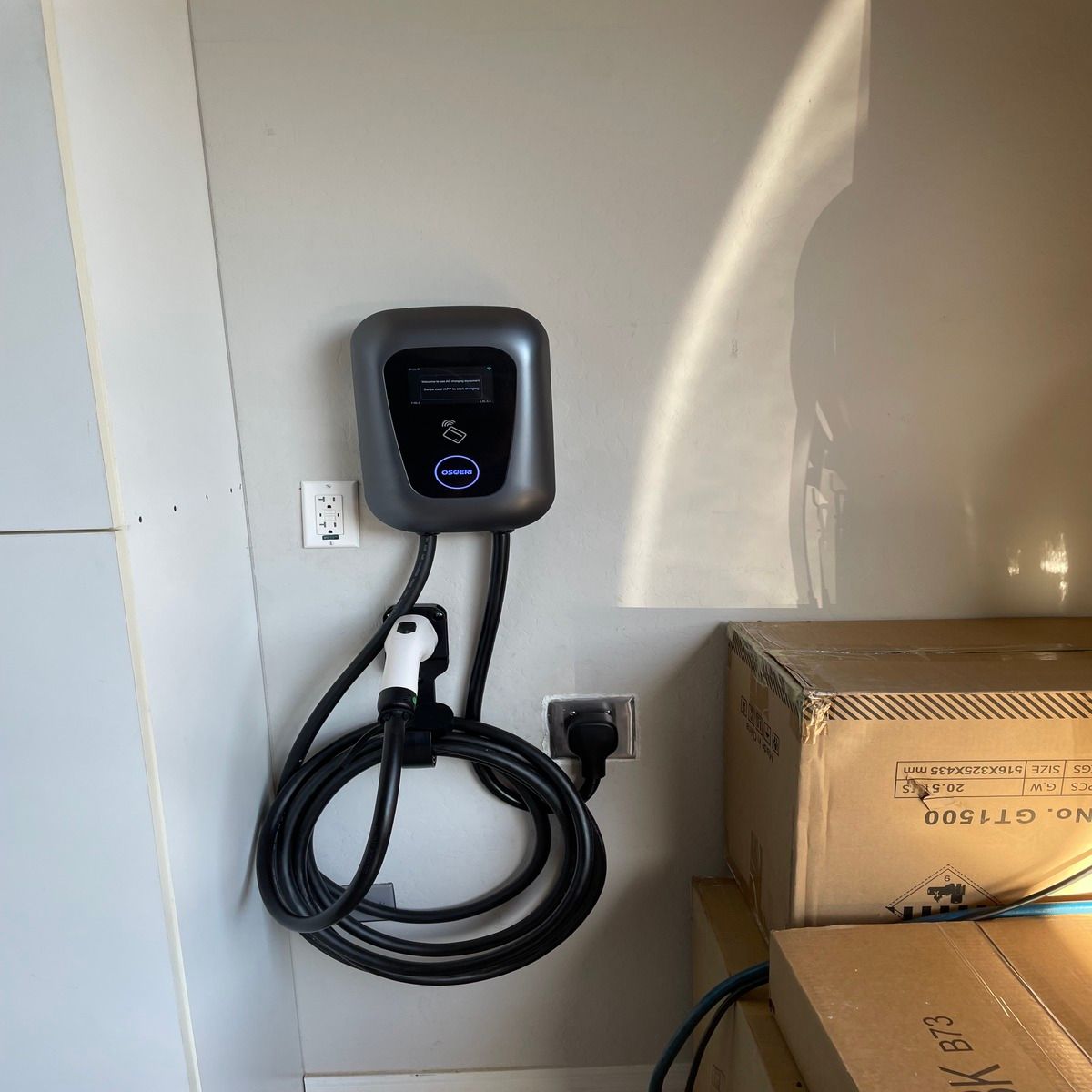 Wall-mounted EV charger with coiled black cable beside cardboard boxes in a garage.