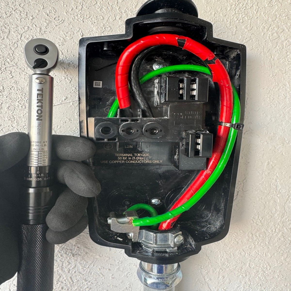 Hand holding a socket wrench beside an open black electrical box with red and green wires.