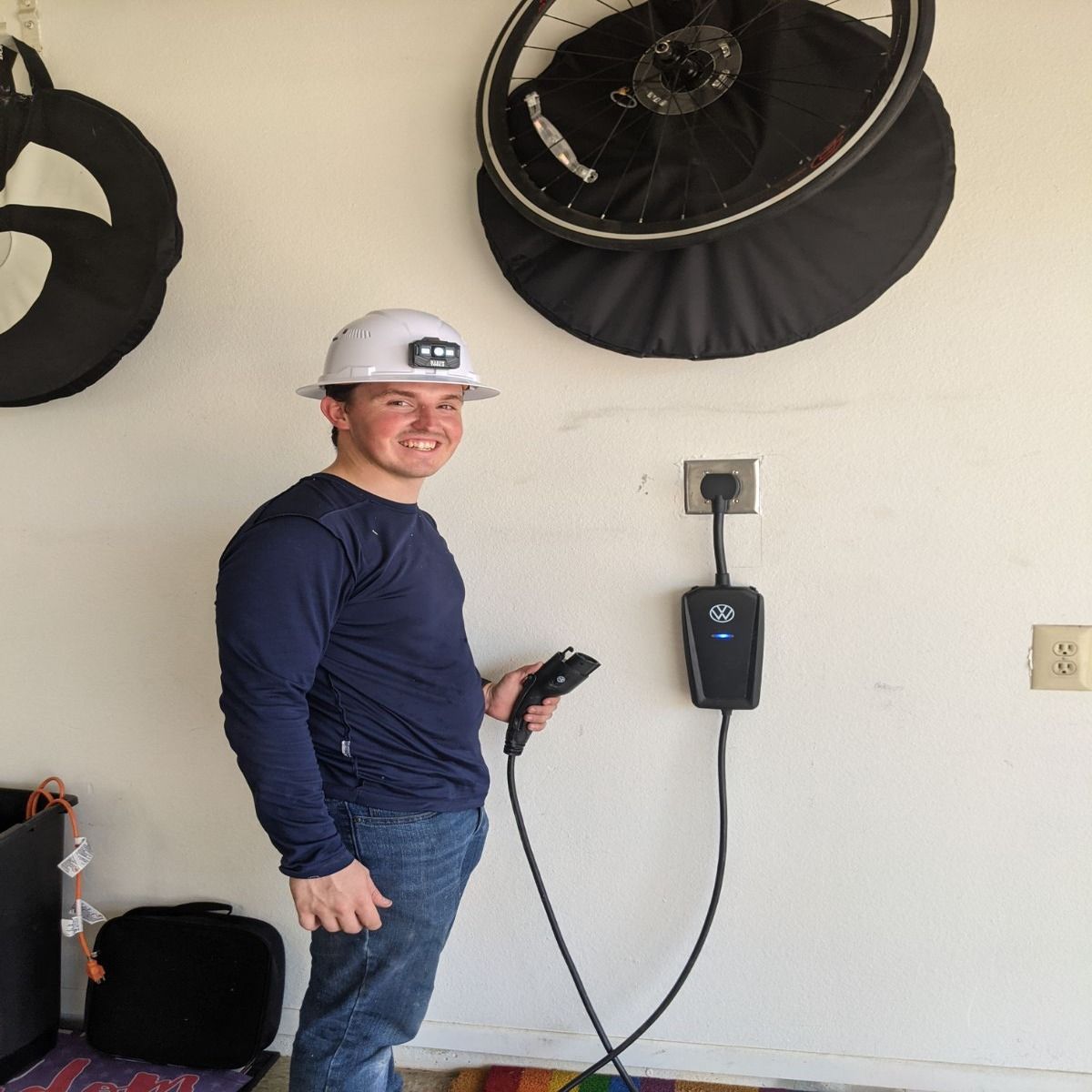 Person in a hard hat smiles while holding a charging cable beside a wall-mounted EV charger in a garage.