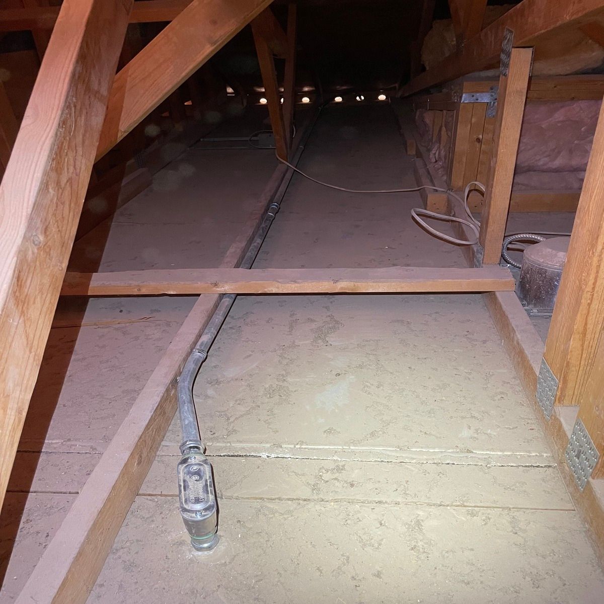 Inside an attic, wooden beams, insulation, and a cable run across the floor and ceiling.