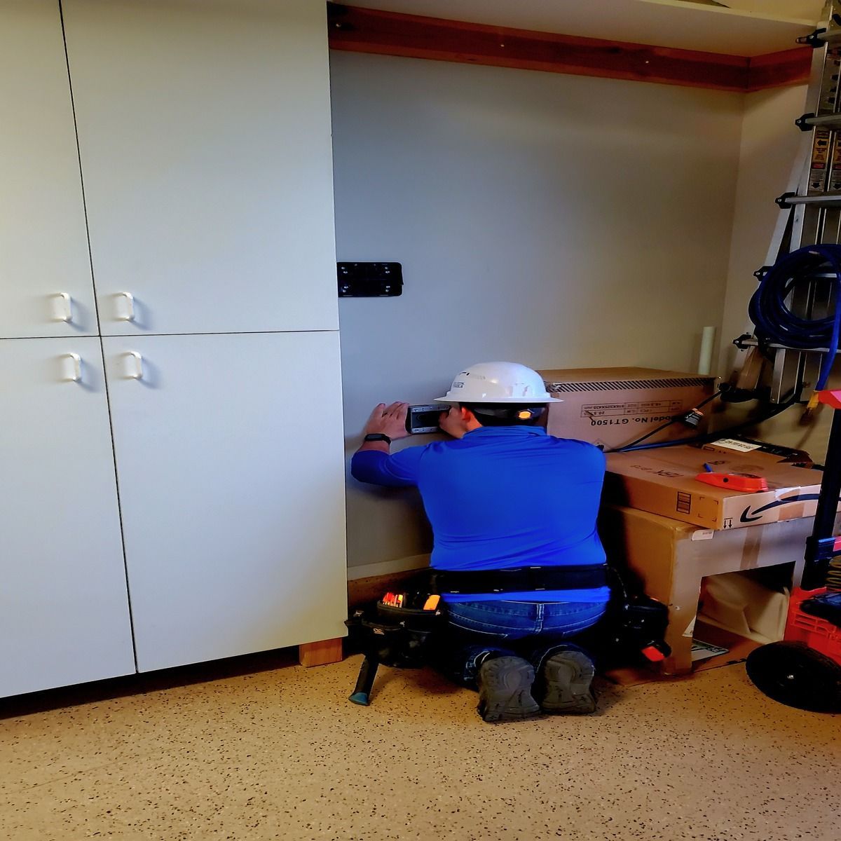 Worker in white hard hat kneels beside cardboard boxes in a garage or storage room.