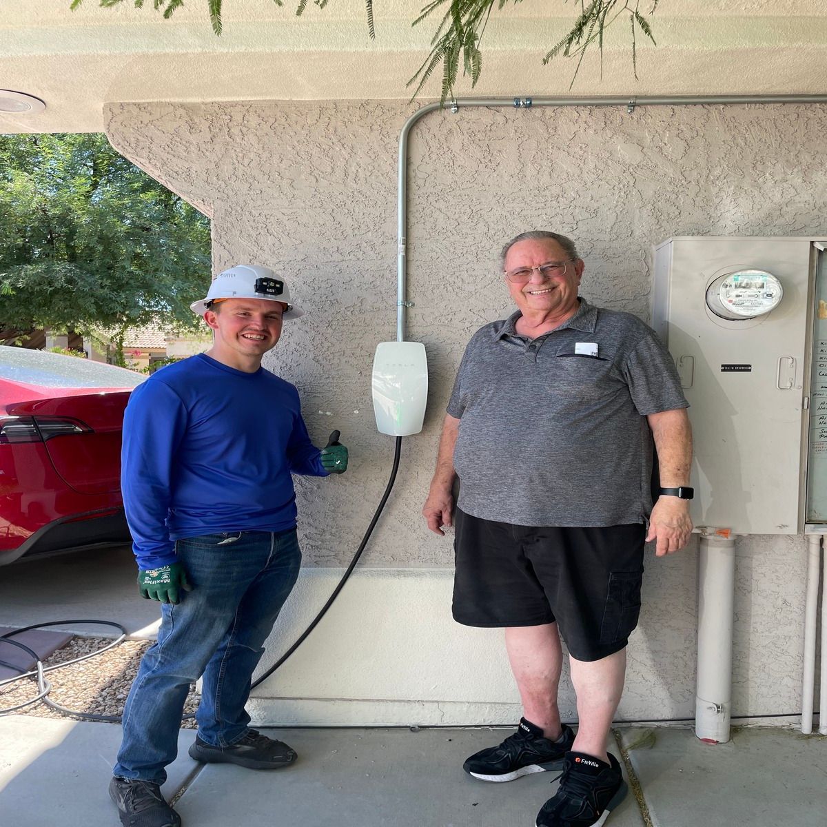 Two men standing by a house exterior, near a red car and utility meter, one smiling at the camera