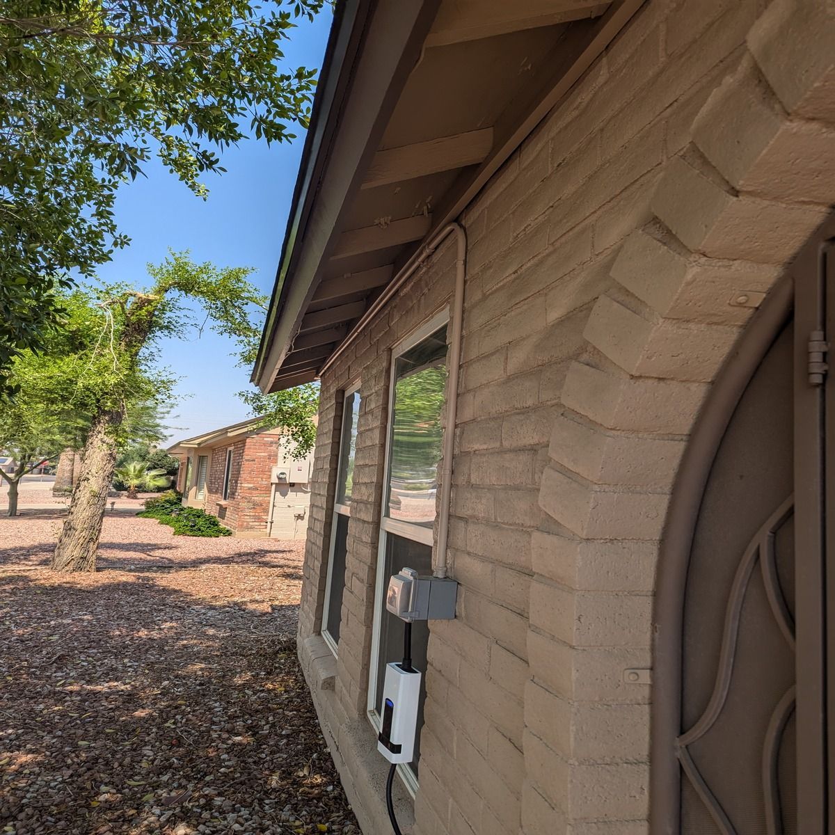 Side view of a tan brick house with a utility meter under the eaves and trees along the yard
