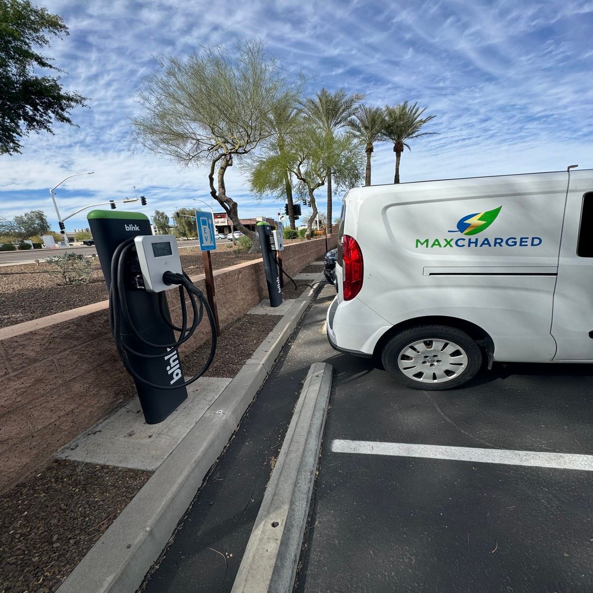 White MaxCharged van plugged into a black EV charger in a parking lot with desert trees and blue sky.