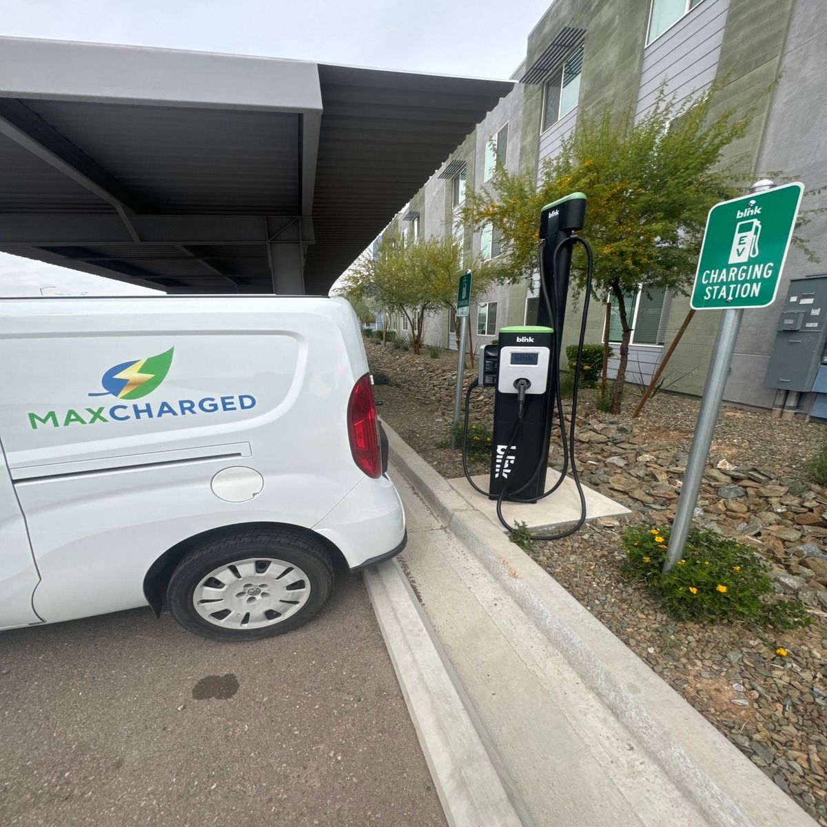White MaxCharged van plugged into an EV charging station under a canopy at curbside.