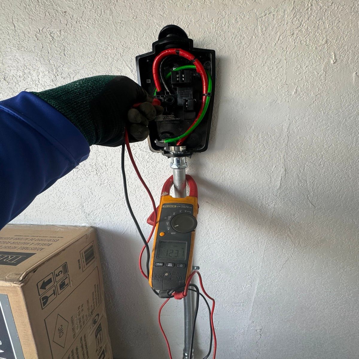 Gloved hand wiring an open electrical outlet box on a wall, with a multimeter hanging below.
