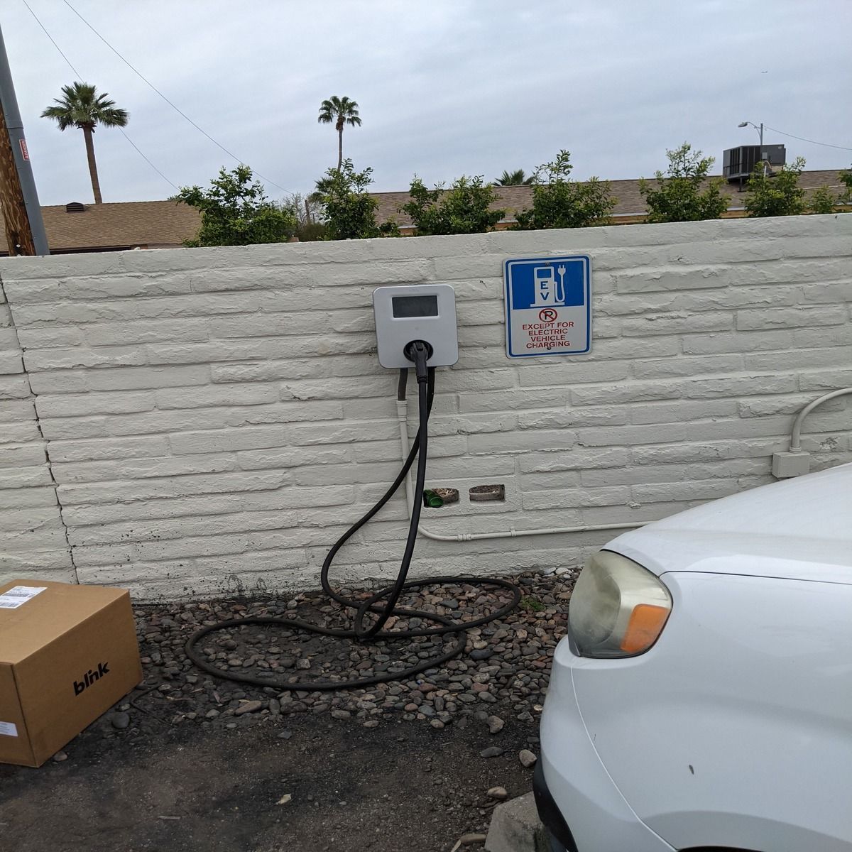Electric vehicle charger on a white wall beside a parked white car, with a cardboard box and dry ground nearby