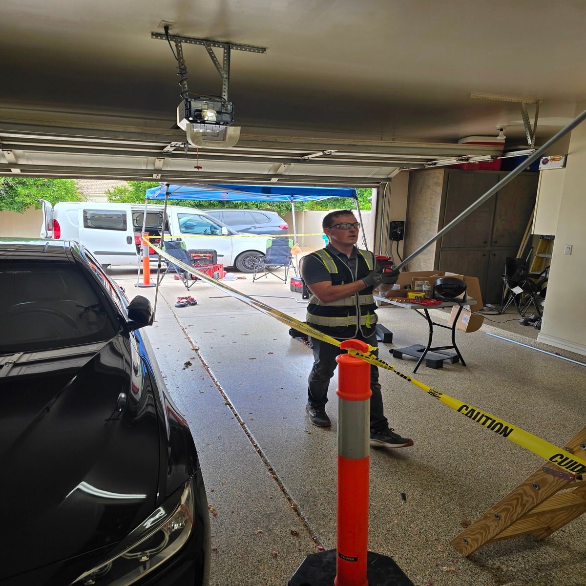 Garage interior with a worker in a safety vest handling caution tape near a parked car and open door.