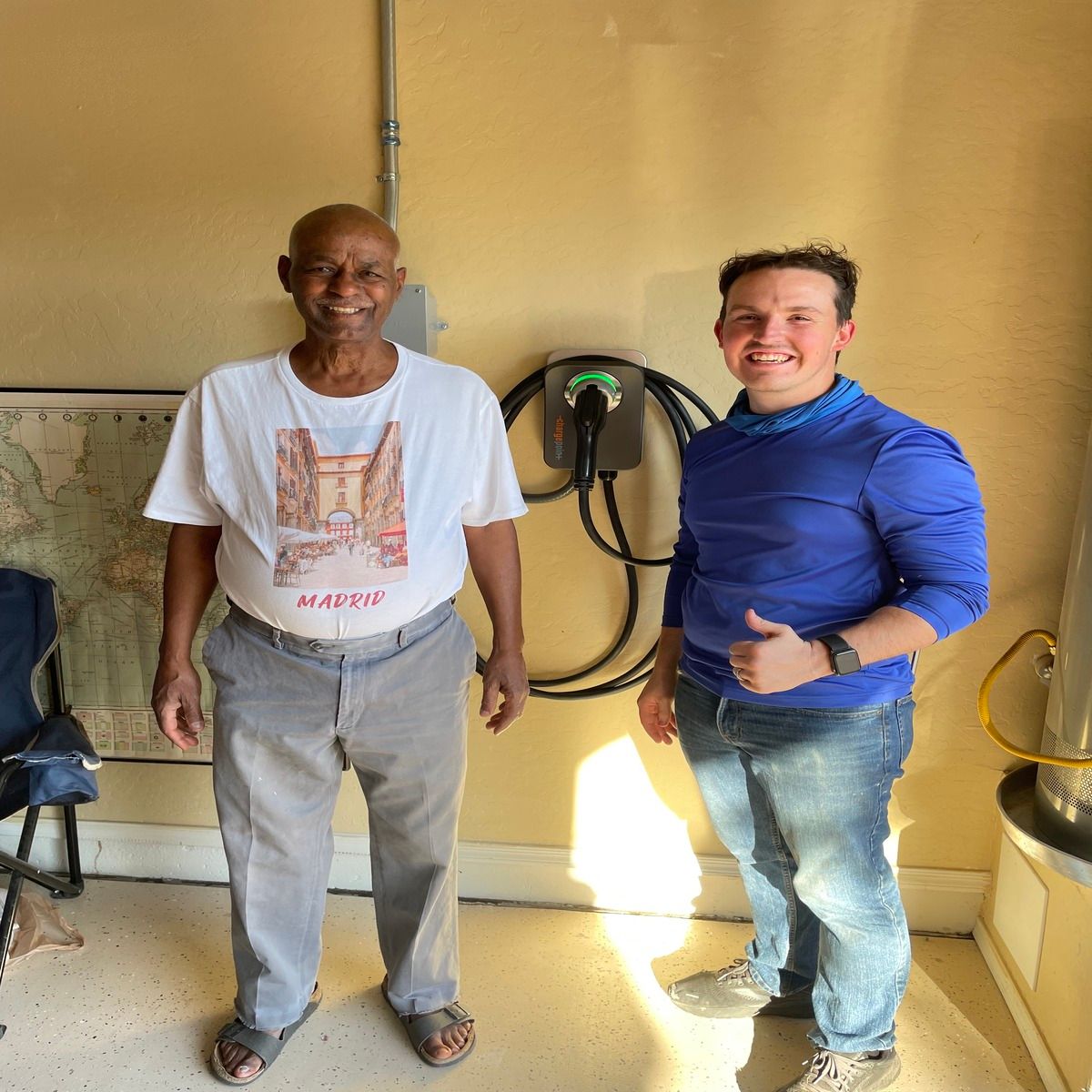 Two people standing indoors by a beige wall, one holding a coiled hose and smiling.