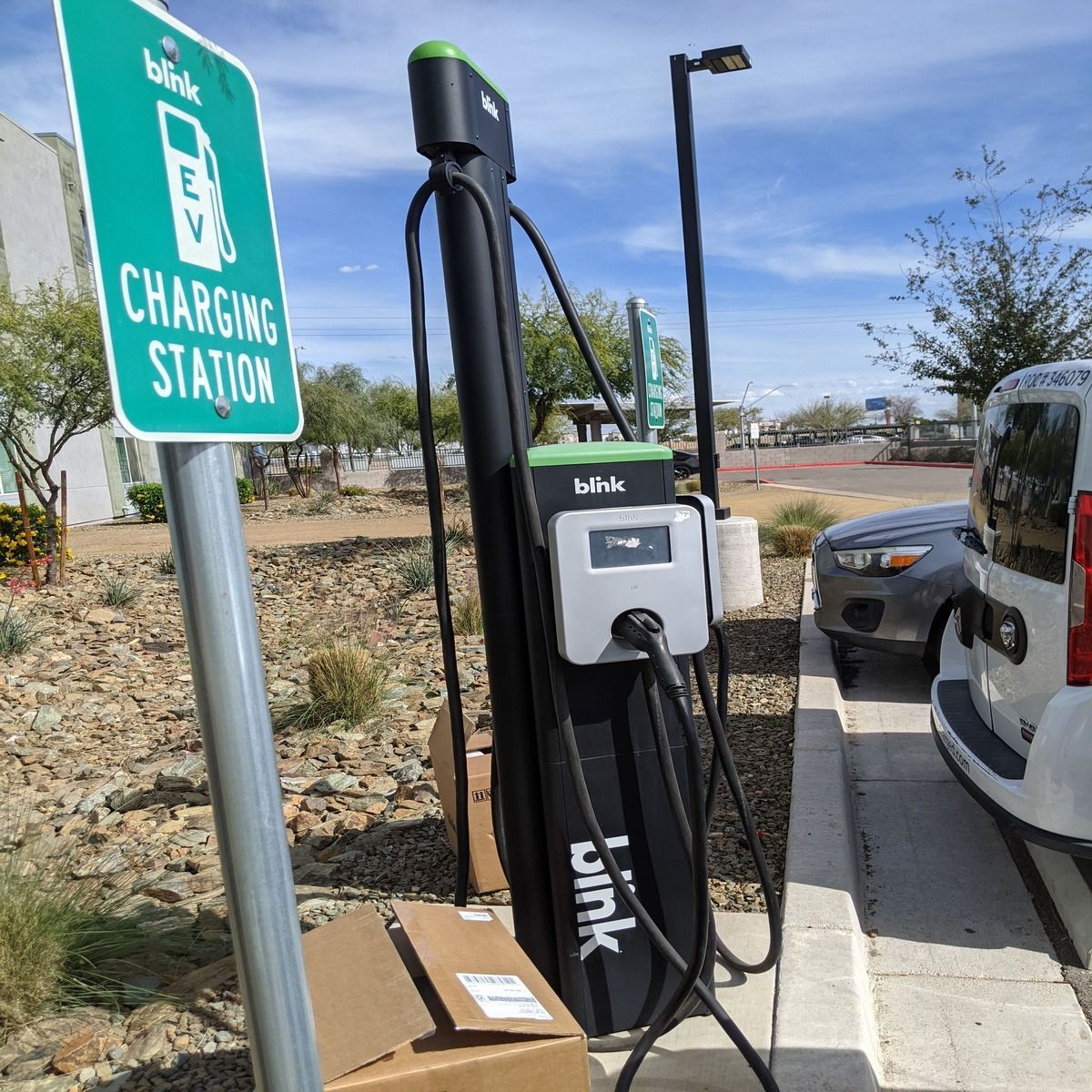 Electric vehicle charging station beside a parking lot, with a white charging unit and green sign.