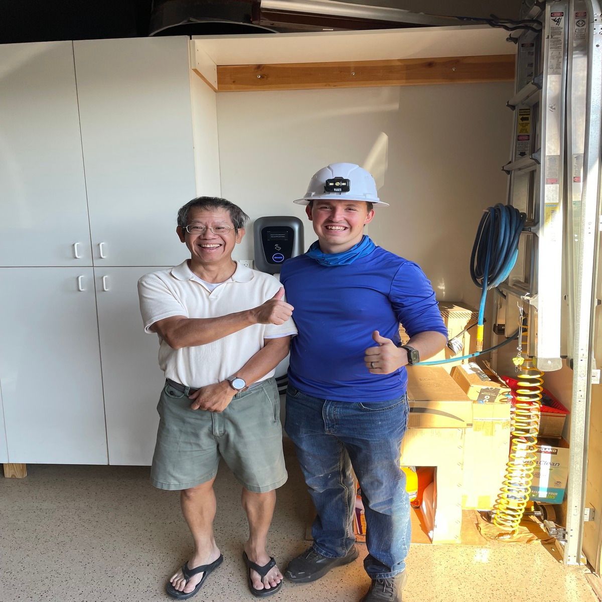 Two people smiling and posing indoors, one in a hard hat near cabinets and tools.