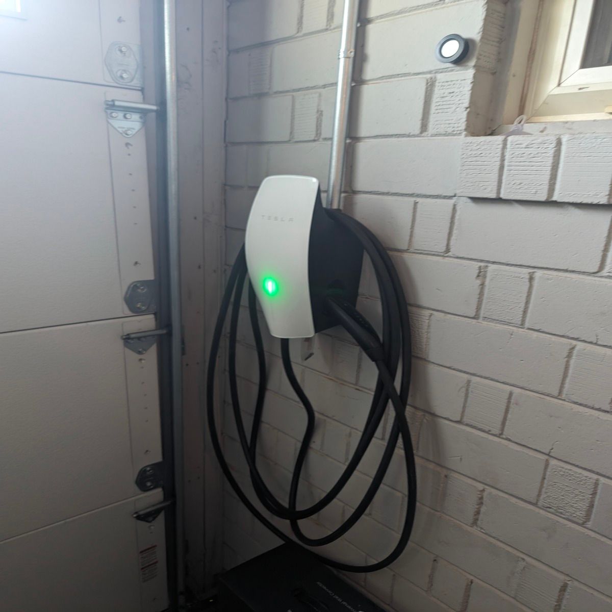 Wall-mounted white EV charger with green light and coiled black cable beside a garage door
