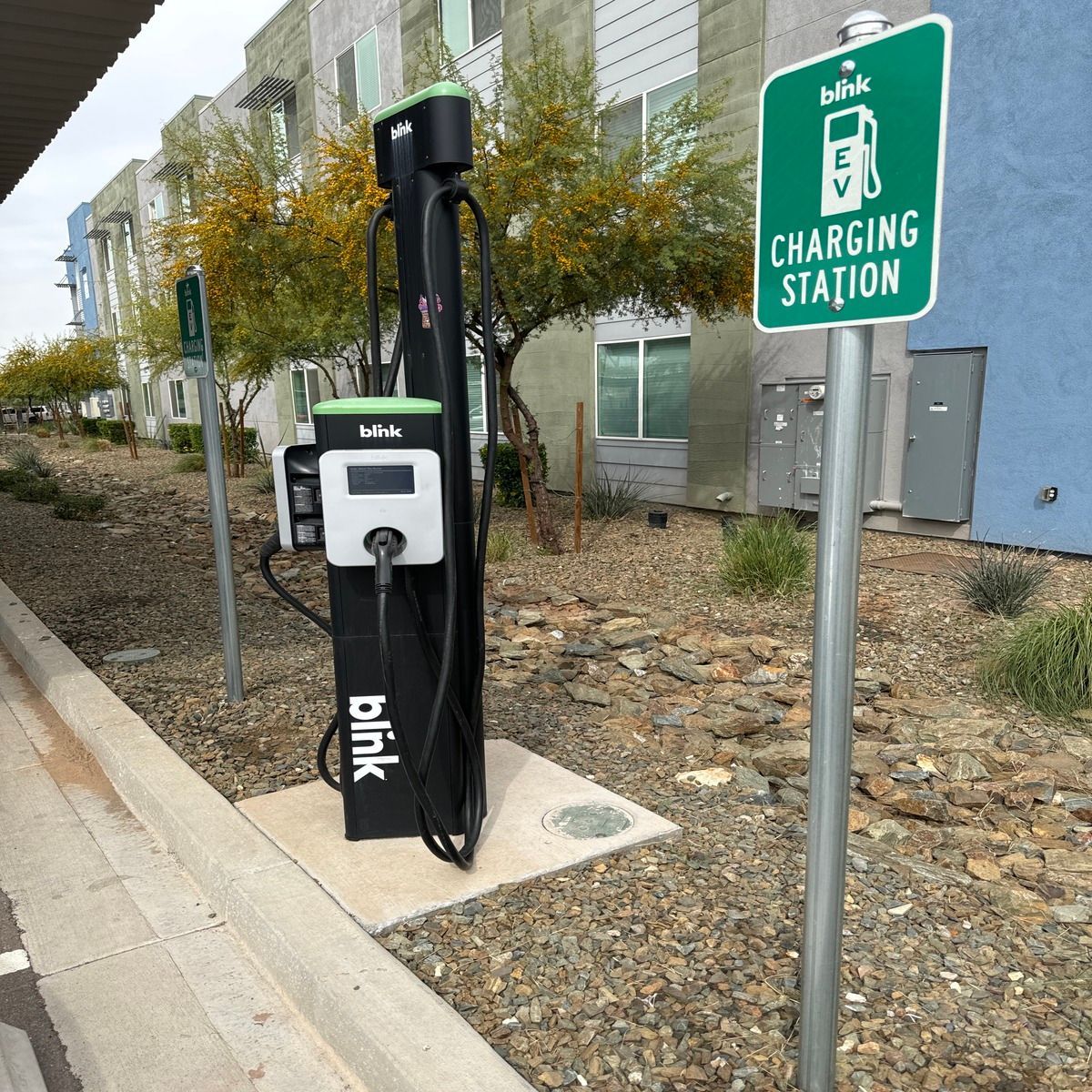 Electric bike charging station on a sidewalk beside a green charging sign and apartment building.