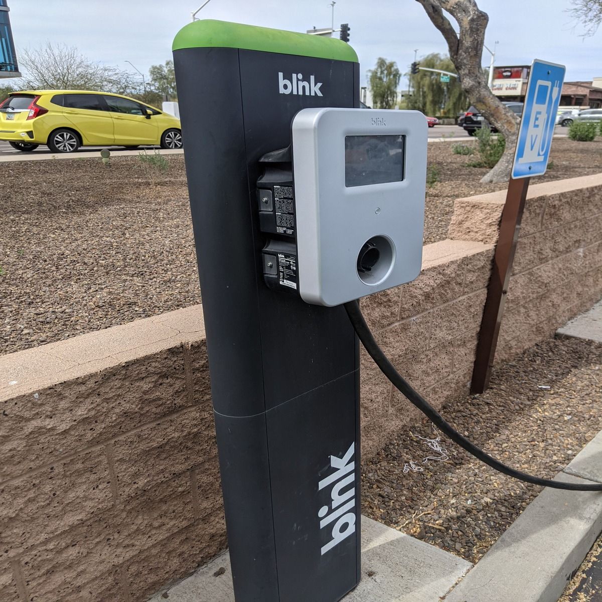 Blink EV charging station on a curbside, black and gray with a green top and connected charging cable.