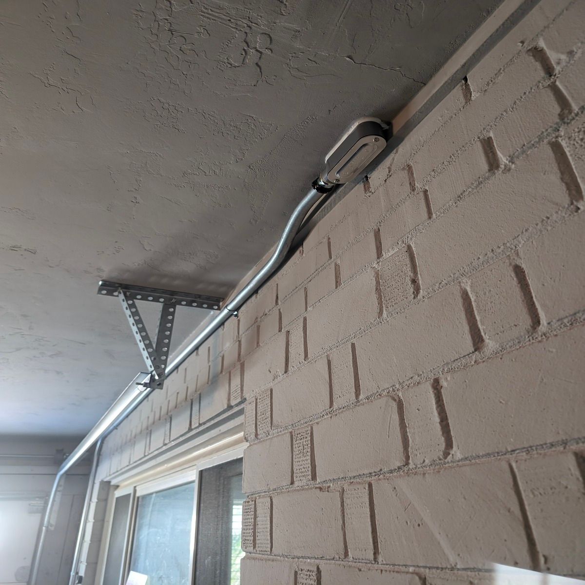 Metal conduit with cable running along an apartment balcony ceiling beside a beige brick wall.