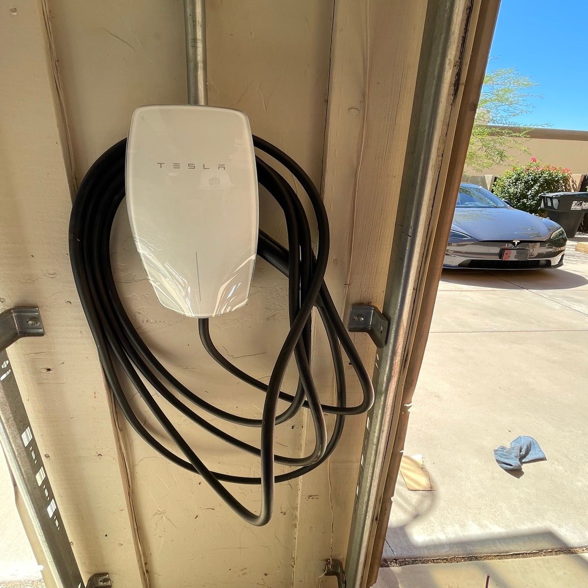 White wall-mounted EV charger with coiled black cable in a garage doorway, sunlight outside