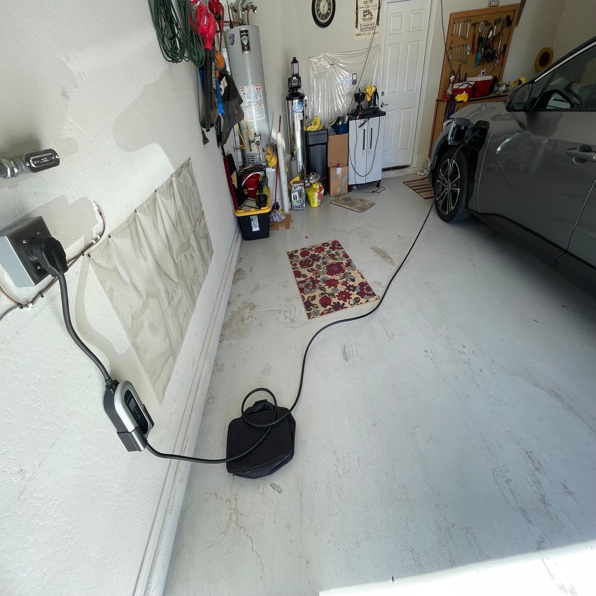 A garage with a gray car, wall-mounted cable reel, floor mat, and cleaning supplies along the back wall.