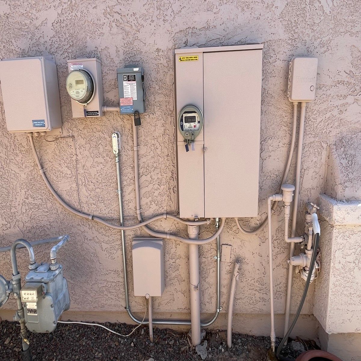 Wall-mounted utility meters and electrical boxes on a stucco exterior wall, with pipes and conduit.
