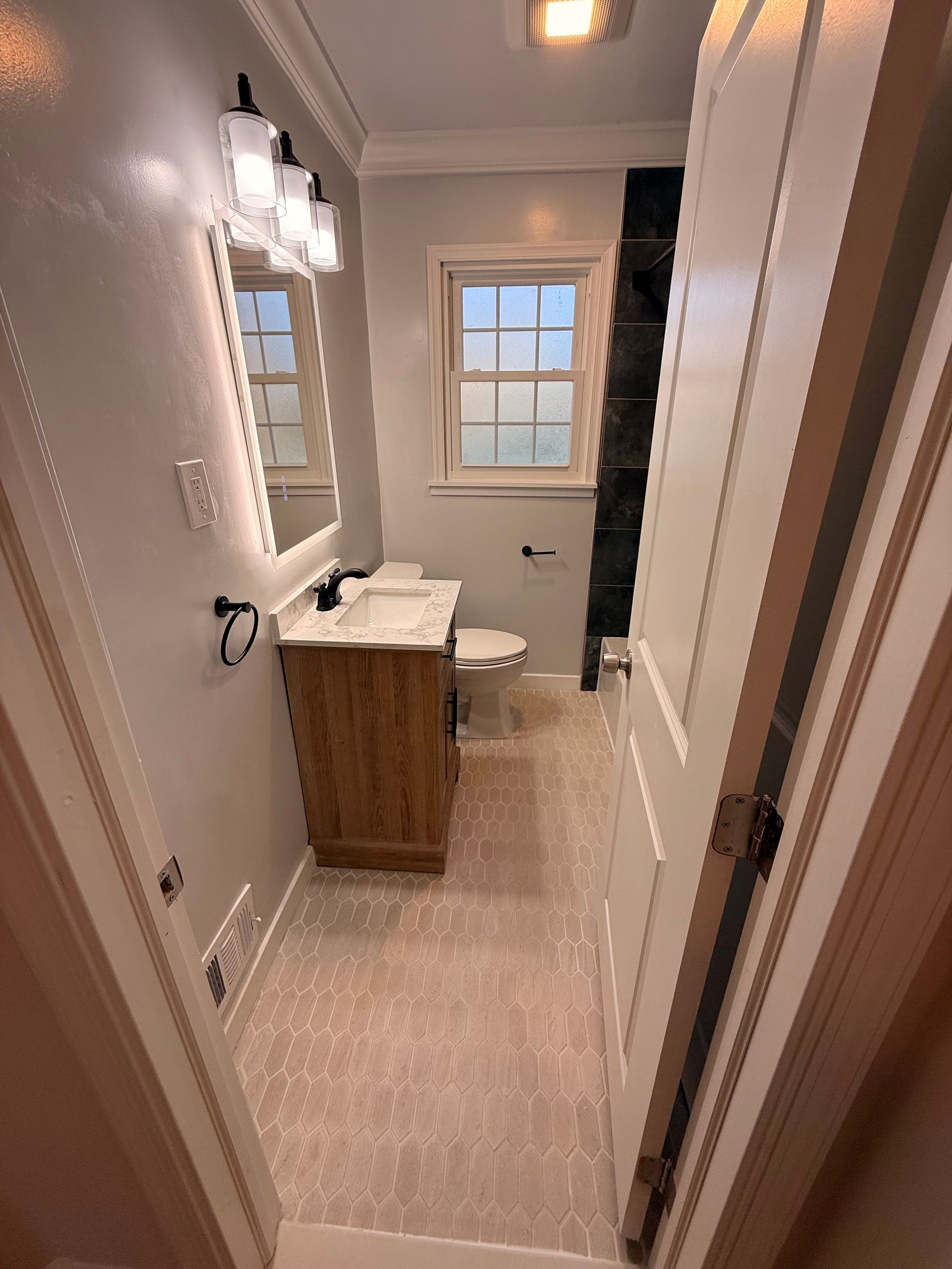Narrow bathroom with vanity, mirror, wall sconce, and window beside a toilet.