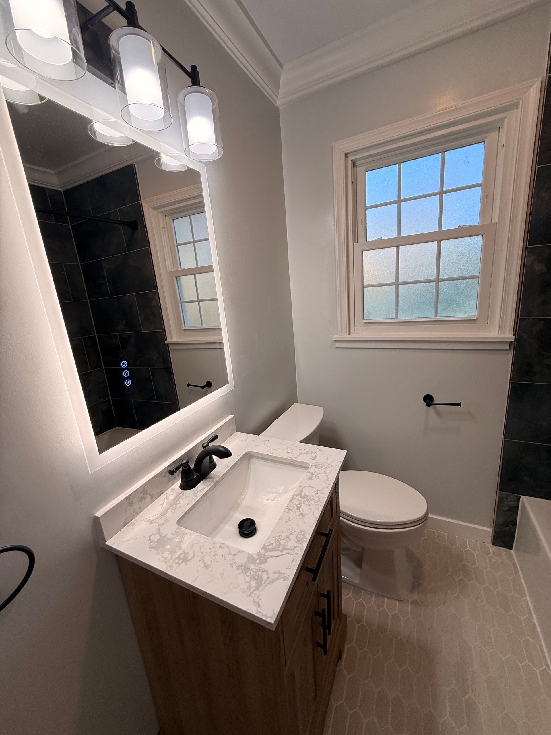 Bright bathroom with white vanity, mirror, toilet, and window, lit by a three-light fixture.