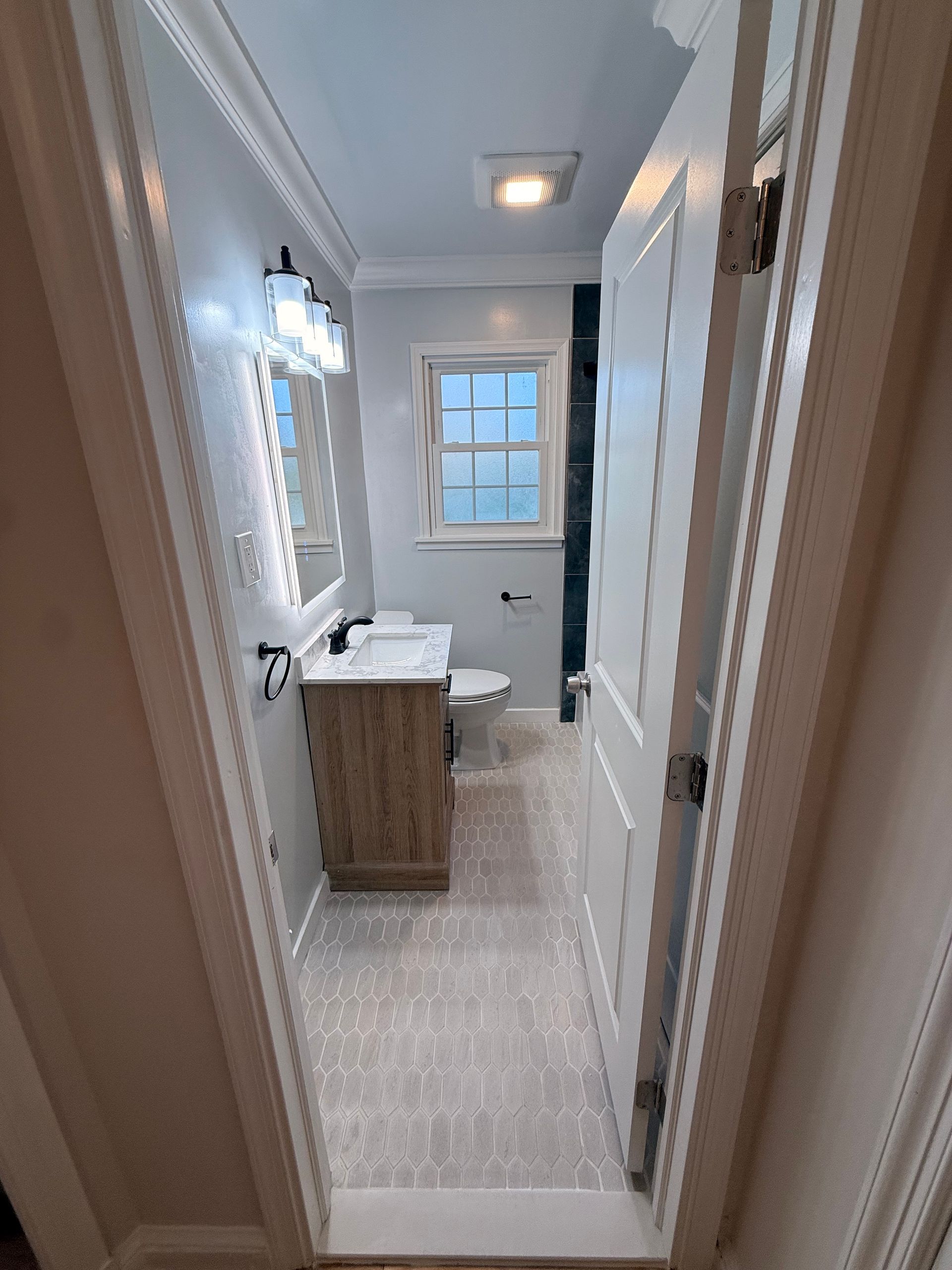 Narrow bathroom with light gray walls, wood vanity, toilet, mirror, and window at the end of the hall.
