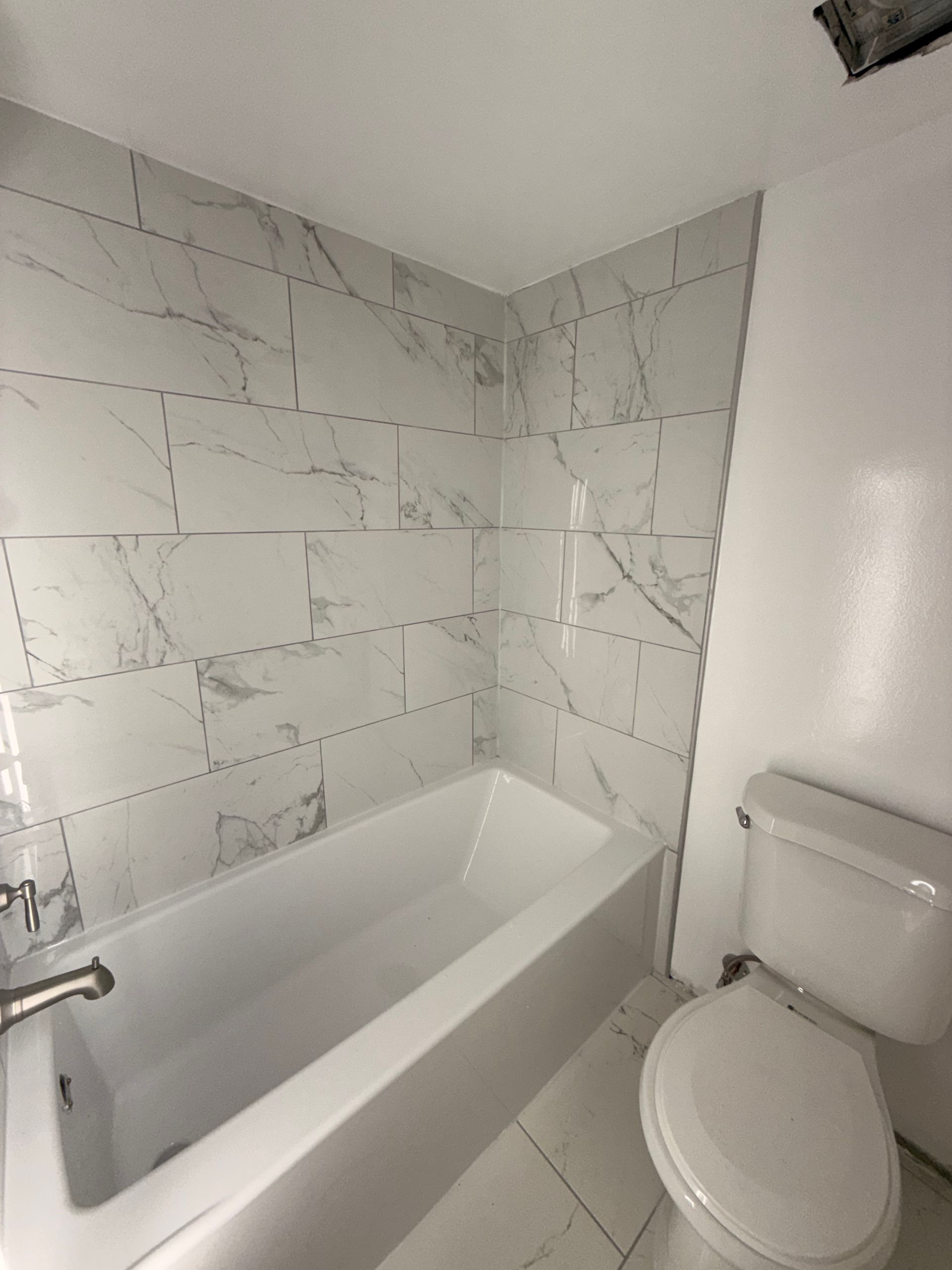 Small white bathroom with a bathtub, marble-look tile walls, and a toilet beside it.