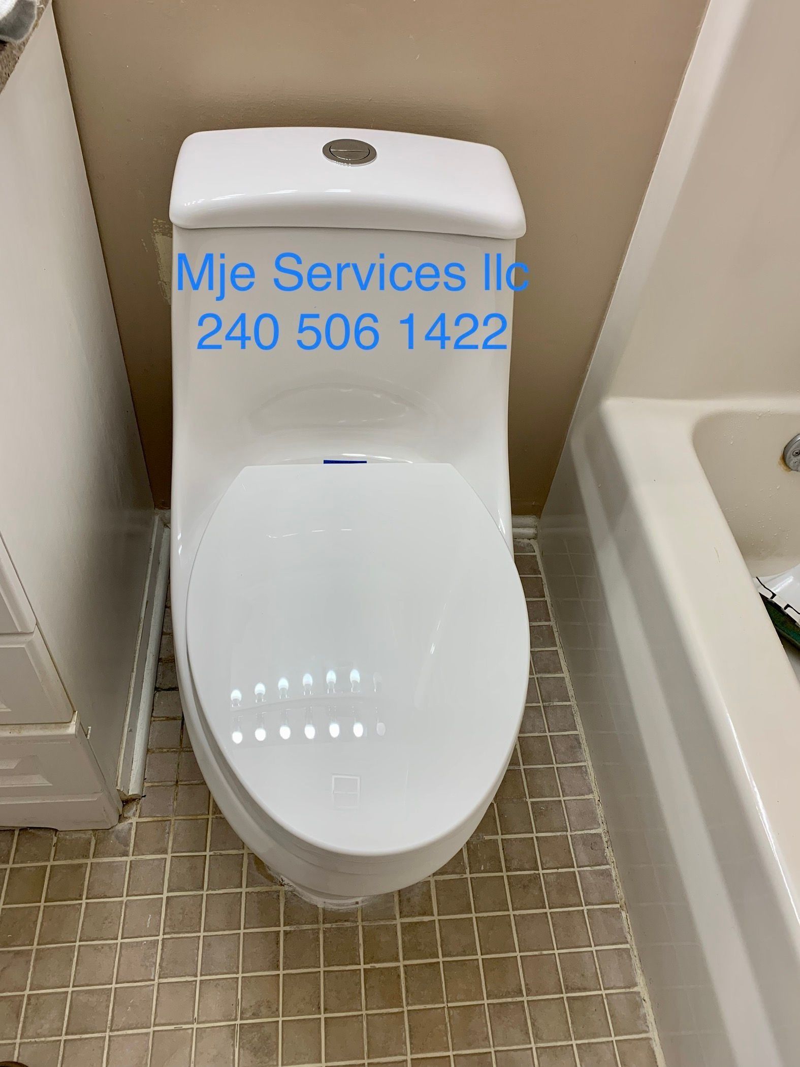 White toilet in a tiled restroom with blue text on the tank reading “Mje Services 240 506 1422”