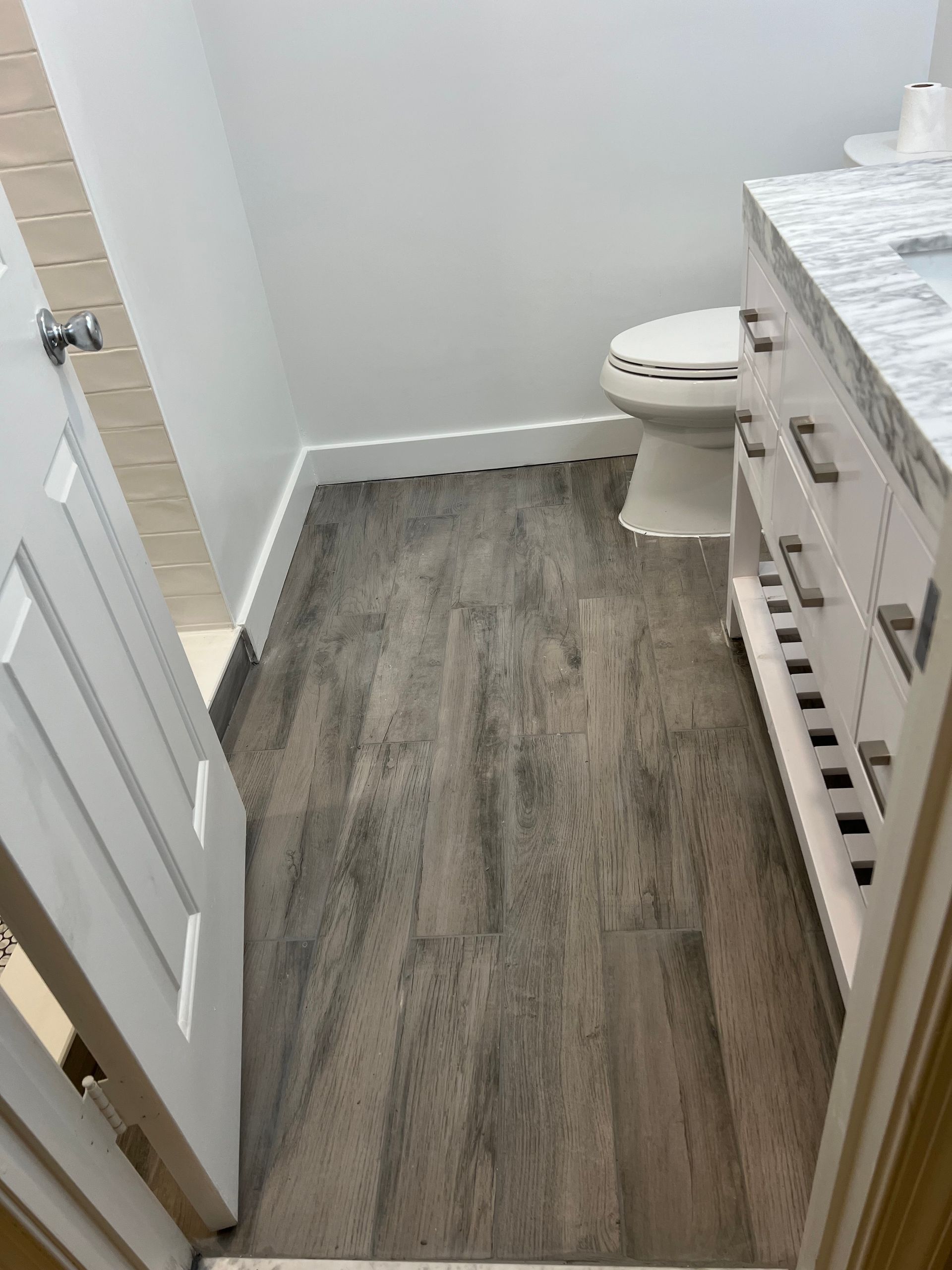 Small bathroom with gray wood-look floor, white toilet, vanity, and open door.