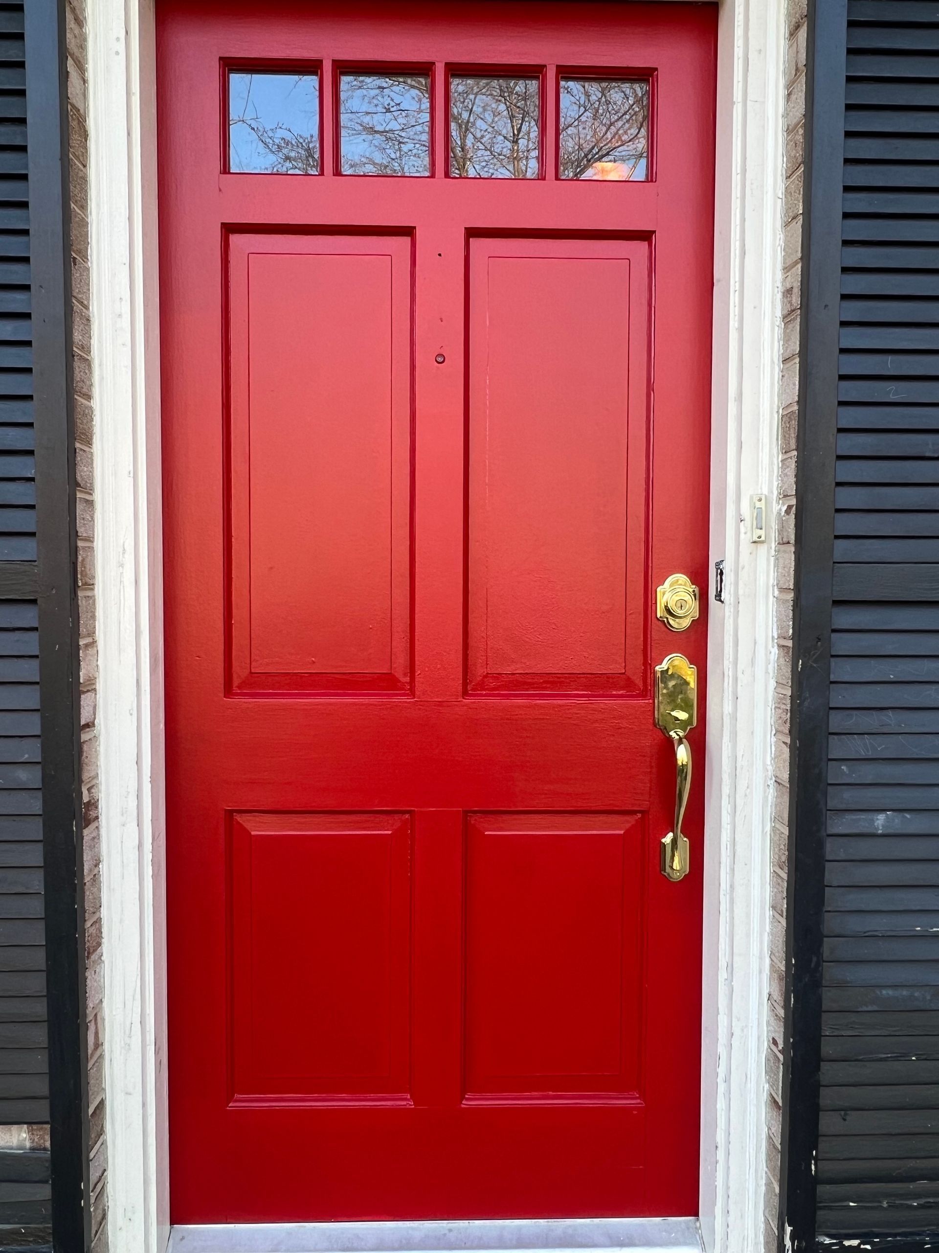Red front door with brass handle and sidelights set in a dark exterior wall.