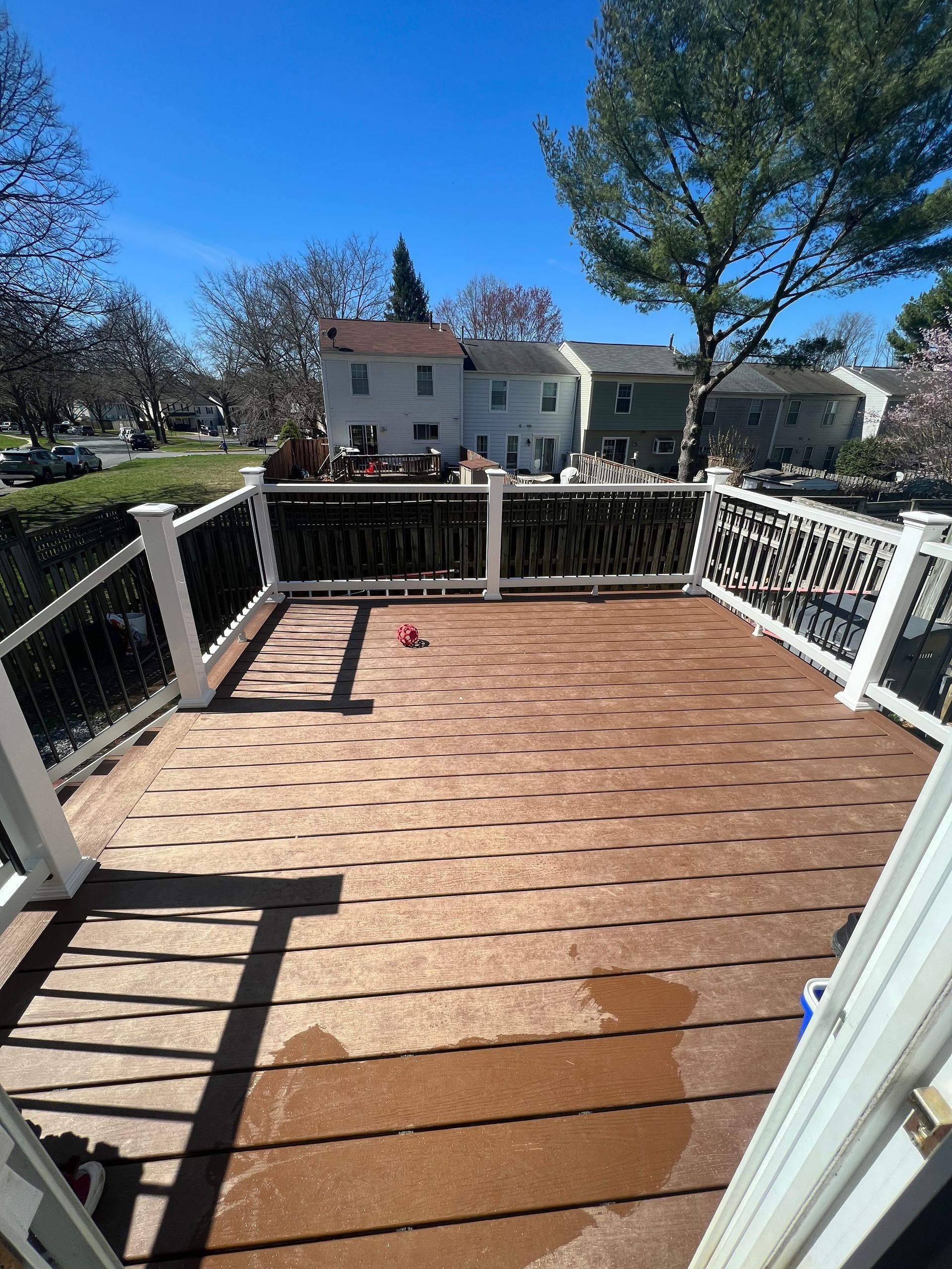 Wooden deck with white railings overlooking a sunny backyard and houses beyond.