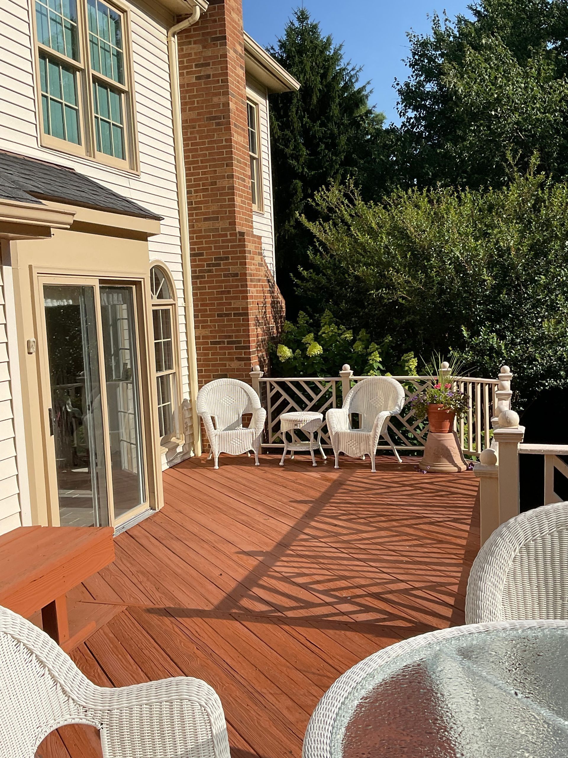 Sunlit wooden deck beside a house, with white patio chairs and a glass table overlooking trees.