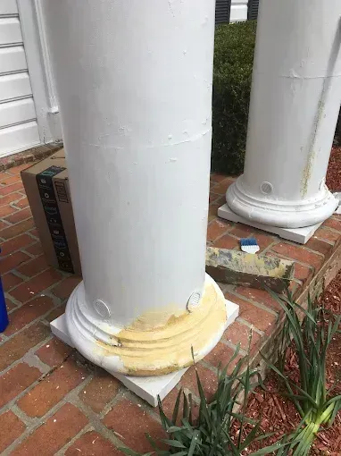 White porch columns with damaged peeling paint and yellowed caulking at the base on a brick patio.