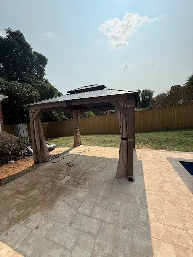 Backyard patio with a beige gazebo canopy beside a pool and wooden fence under a bright sky