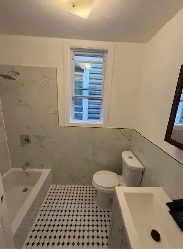 Small bathroom with white and gray tile, a toilet, tub, sink, and a narrow window.