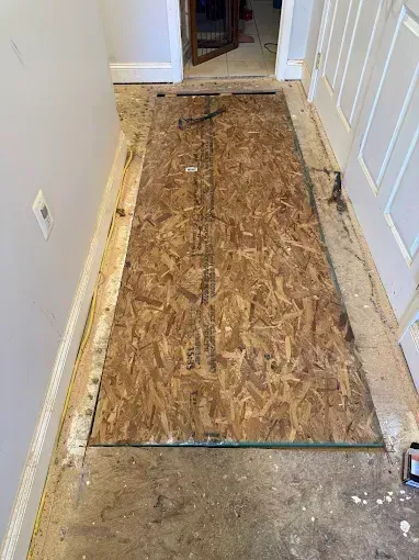 Narrow hallway with a plywood subfloor patch over damaged flooring, leading to a doorway.