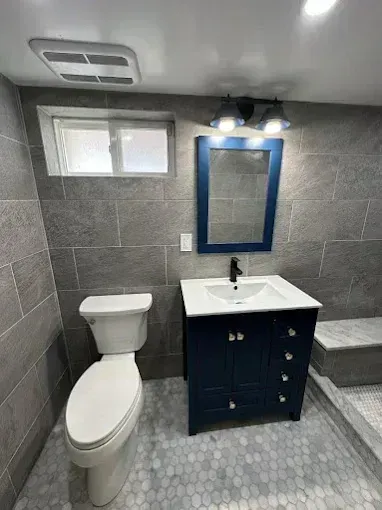 Small gray-tiled bathroom with a toilet, vanity sink, blue-framed mirror, and step-up shower.