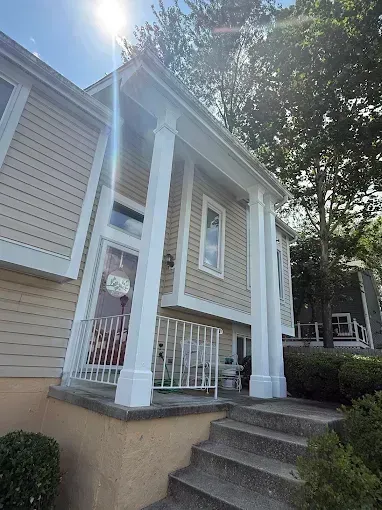 Front porch of a beige house with white columns, railing, and steps in bright sunlight