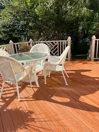 White patio chairs and glass table on a sunlit wooden deck with railing and trees beyond